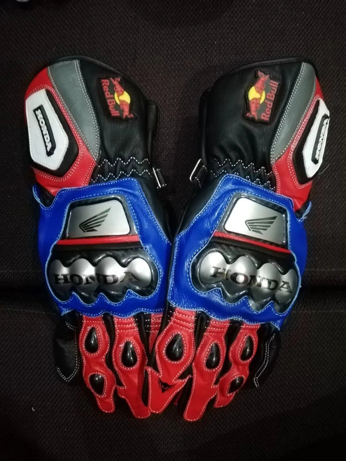Honda and Red Bull branded leather motorcycle racing gloves with blue and red panels and metallic knuckle armor.