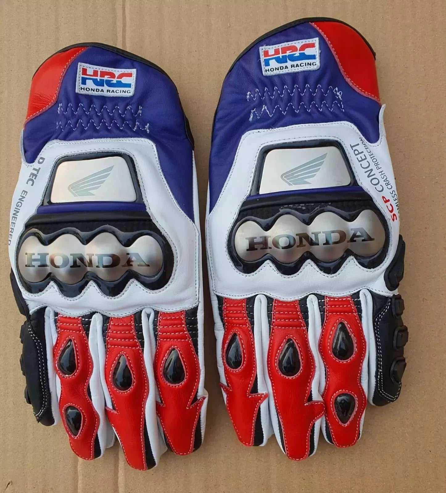 Honda HRC leather motorcycle racing gloves in red, white, and blue with metallic knuckle armor.