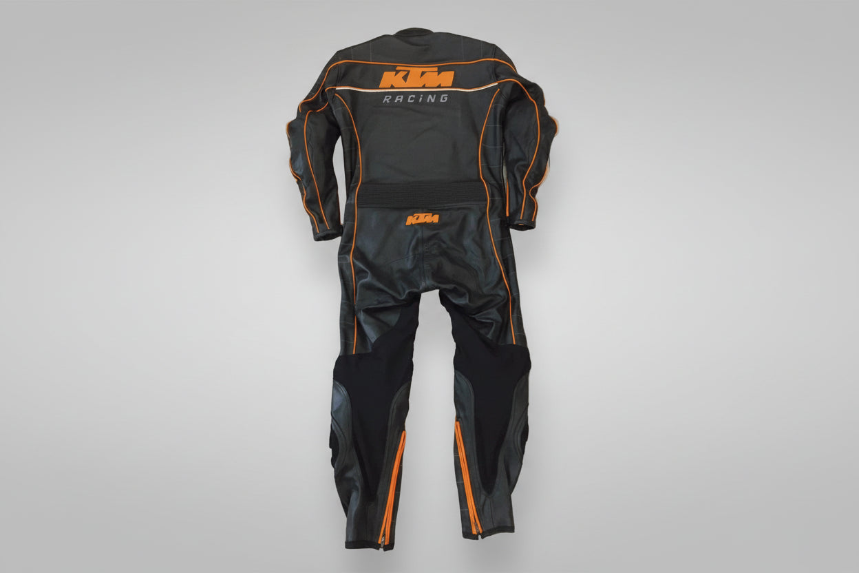 Detailed view of the KTM Racing chest logo, orange technical piping, and accordion stretch panels on a professional racing suit.